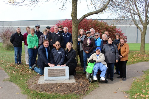 Image of a group of people attending the ceremony dedicating the plaque for Paul R. Hoffman with a walking path on both sides of the group, a tree turning red for fall, and an industrial building in the far background.